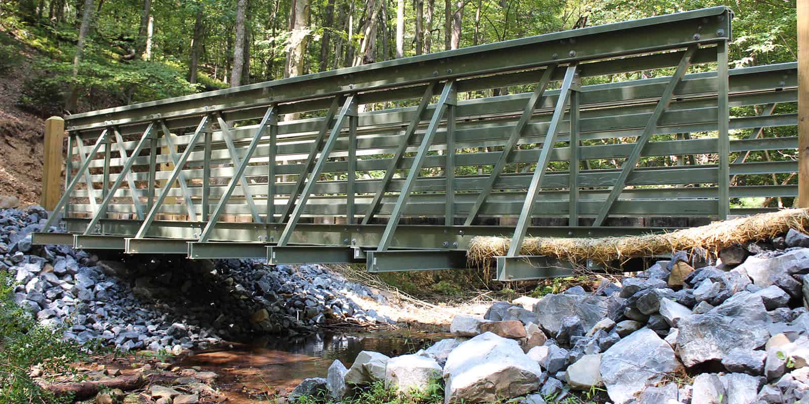 Green ReadySpan pedestrian bridge in a wooded area spanning a creek with rocks.