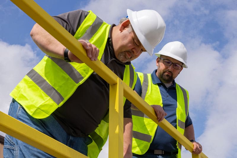 Two contsruction workers in vests and hard hats looking over yellow railing