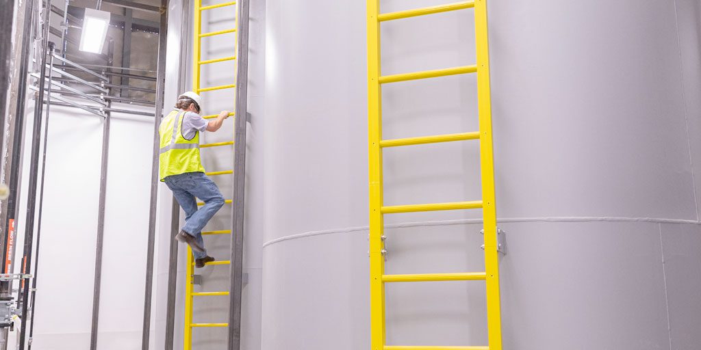 Man in safety gear climbing fixed ladder in industrial setting.